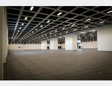 Große, leere Messehalle mit einer Decke aus quadratischen Gitterelementen, hellen Leuchtstofflampen und mehreren weißen Tragpfeilern. Der Boden ist gefliest, und die Wände sind schlicht weiß mit wenigen Türen und Fenstern.