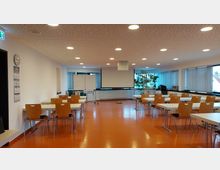 Ein moderner Seminarraum mit orangefarbenem Boden, mehreren rechteckigen Tischen und Holzstühlen. Im Hintergrund befinden sich eine Leinwand, Flipcharts, ein Projektor, Pflanzen und große Fenster mit Jalousien.