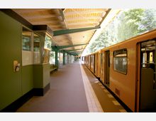 U-Bahn-Haltestelle mit überdachtem Bahnsteig, grünen Stützpfeilern und gelbem U-Bahn-Zug auf der rechten Seite. Im Hintergrund sind Bäume und weitere Bahnhofselemente sichtbar.