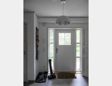 Eingangsbereich eines Hauses mit einer weißen Tür, die von zwei schmalen Fenstern flankiert ist. Im Vordergrund stehen Schuhe und Accessoires auf einer Matte und Ablage, darüber hängt eine schlichte, halbrunde Deckenleuchte.