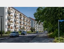 Wohnstraße mit mehrstöckigen Wohnhäusern auf der linken Seite, gekennzeichnet durch zahlreiche Balkone mit Pflanzenschmuck. Rechts ist eine Baumreihe zu sehen, die Schatten auf den Gehweg und die Fahrbahn wirft. Straßenschild „Birkenweg“ sichtbar, geparkte Autos entlang der Straße.