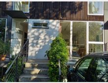 Moderne Wohnhausfassade mit einer Eingangstür links, umgeben von Pflanzen und Glasfenstern, die teilweise den Innenraum zeigen. Eine kleine Treppe führt zum Eingang, daneben ist ein dunkles Auto geparkt.