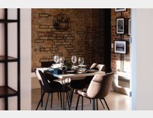 Essbereich in einem industriellen Stil mit freiliegenden Backsteinmauern. Ein Esstisch, gedeckt mit Geschirr und Weingläsern, ist umgeben von gepolsterten Stühlen; an der Wand hängen gerahmte Schwarz-Weiß-Bilder.