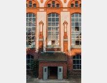 Fassade eines historischen Backsteingebäudes mit großen Bogenfenstern und verzierten weißen Putzdetails. Im Vordergrund ein kleineres Vordach mit grünlichen Doppeltüren und Dachziegeln.