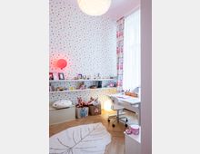 Kinderzimmer mit bunten Punkten an der Wand und einem hellen Holzfußboden. Zwei Regale mit Spielzeug und Dekorationen, ein Schreibtisch mit weißem Stuhl und eine große Fensterfront mit gemusterten Vorhängen dominieren den Raum; ein Teppich in Blattform liegt auf dem Boden.