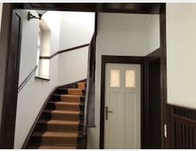 Treppenhaus in einem Wohngebäude mit einer Holztreppe, die mit einem braunen Teppichläufer belegt ist. Rechts daneben befindet sich eine weiße Tür mit Milchglaseinsätzen, und das Geländer sowie die Türrahmen sind dunkelbraun lackiert.