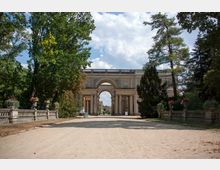 Sandweg, der von Bäumen und Balustraden gesäumt ist, führt zu einem historischen Torbau mit einem großen Bogendurchgang und dekorativen Säulen; im Hintergrund ist weiteres klassizistisches Gebäude erkennbar. Der Himmel ist leicht bewölkt.