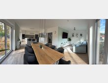 Offener Wohn- und Essbereich mit hellem Holzboden, einem großen Esstisch aus Holz mit schwarzen Stühlen und einer modernen Küche im Hintergrund. Im angrenzenden Wohnzimmer stehen eine helle Sofalandschaft, ein Wandfernseher und Dekorationen; große Fenster lassen viel Tageslicht herein.