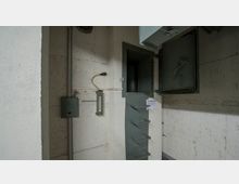 Betonraum mit technischen Einrichtungen, darunter eine Überdruckmesseinrichtung und eine kleine Metallleiter, die zu einer schmalen Türöffnung führt. An der Wand befinden sich Kabel und ein graues Kästchen, die Teil der technischen Installation sind.