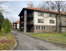 Mehrstöckiges Gebäude mit hellen Putzfassaden und Steinelementen, roten Ziegeldächern und Balkonen aus dunklem Holz. Im Vordergrund ein gepflasterter Weg, Rasenflächen, Bäume und Büsche, daneben ein orangefarbener Mülleimer und eine grüne Bank.