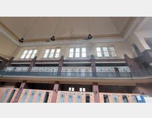 Ein historischer Innenraum mit mehreren Etagen, geprägt von Backsteinwänden, hohen Fenstern mit teils buntem Glas und großen Lampen, die von der Decke hängen. Die Architektur zeigt ein symmetrisches Layout mit Metallgeländern und Türen auf beiden Ebenen.