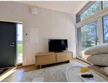 Heller Wohnbereich mit weißer Holzvertäfelung und hoher Decke, Fernseher auf einem niedrigen Sideboard sowie hellem Teppichboden. Große Fensterfront und Glastür geben den Blick auf Bäume und eine Grünfläche im Außenbereich frei.