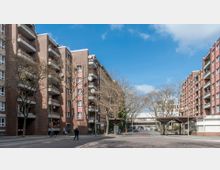 Eine Wohnstraße mit mehrstöckigen Backsteingebäuden auf beiden Seiten, die Balkone mit Satellitenschüsseln und Pflanzen haben. Im Hintergrund sind eine Brücke und Bäume zu sehen, während das Straßenpflaster und eine überdachte Bushaltestelle den Vordergrund bestimmen.