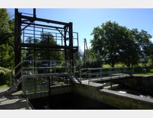 Wasserschleuse mit metallenen Geländern und einer Brücke, umgeben von Bäumen und Grünflächen. Im Hintergrund sind ein Auto, ein Fluss und blauer Himmel sichtbar.