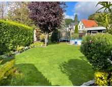 Ein gepflegter Garten mit einer großen Rasenfläche, umgeben von Hecken, Sträuchern und Blumen. Im Hintergrund sind ein Baum mit rotem Laub, ein Pool mit Leiter, ein Trampolin sowie Häuser mit roten und grauen Dächern sichtbar.