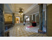 Großzügiges historisches Foyer mit Marmorsäulen, einer Treppe mit Balustrade, einer Marmorbodenfläche im Schachbrettmuster und einem Kronleuchter an der Decke. Im Hintergrund führen Gänge und Treppen zu weiteren Bereichen des Gebäudes.