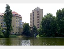 Wohngebäude in einer urbanen Umgebung, mit einem niedrigen Altbau mit roten Dächern und großen Fenstern neben einem hohen, hellbeigen Plattenbau; im Vordergrund ein kleiner Fluss oder Teich, umgeben von dichtem Grün. Eine gemauerte Brücke mit hübschen Säulen führt durch das Wasser.