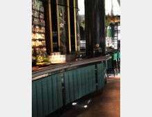 Eine stilvolle Bar mit gepolsterter, türkisfarbener Front und schwarzer Thekenplatte. Im Hintergrund befinden sich Regale mit Flaschen, dunkle Säulen, Pflanzen- und Blumenmotive an den Wänden sowie dekorative Beleuchtung.