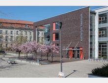 Moderne Backsteinfassade des „auditorium maximum“ mit roten und gläsernen Elementen, flankiert von blühenden Kirschbäumen auf einem gepflasterten Platz. Im Hintergrund ist ein historisches Gebäude mit roten Dachziegeln zu sehen.