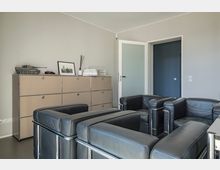 Wohnzimmer mit modernem Design, darin schwarze Ledersessel und ein beiger Metallschrank. Auf dem Schrank stehen ein Modellpanzer, ein gerahmtes Schwarz-Weiß-Foto und Dekorationsgegenstände. Im Hintergrund sind eine Glastür und eine farbige Wand zu sehen.