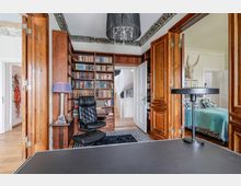 Holzbibliothek mit Bücherregalen an einer Wand, einem schwarzen Ledersessel und einem dunklen Schreibtisch im Vordergrund. Geöffnete doppelte Holzflügeltüren führen in ein angrenzendes Schlafzimmer mit einem Bett und türkisfarbener Bettdecke.