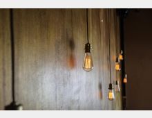 Nahaufnahme von mehreren hängenden Glühbirnen mit sichtbaren Glühfäden vor einer Holz- oder braunen Wand, die eine warme Beleuchtung erzeugen. Die Glühbirnen sind in einfacher Halterung an dünnen Kabeln befestigt.