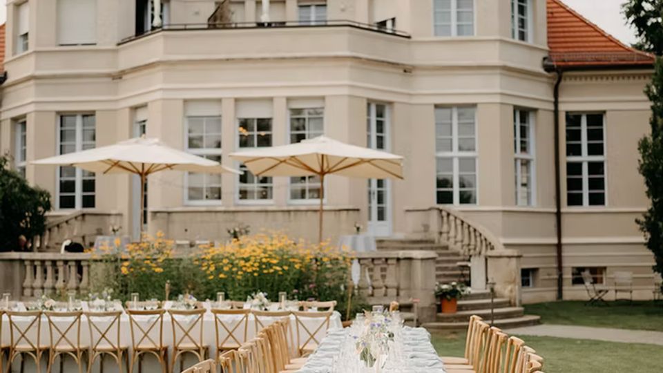 Großer Garten vor einer eleganten Villa mit hellem Putz, vielen hohen Fenstern und rotem Ziegeldach. Auf dem Rasen stehen lange, weiß gedeckte Tafeln mit Holzstühlen, daneben Sonnenschirme, Blumenbeete und eine Steinterrasse mit Treppen.
