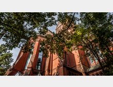Backstein-Kirche mit neugotischen Architekturmerkmalen, darunter Spitzbögen und hohe Fenster, teilweise verdeckt durch Baumäste und Laub. Die Turmspitze mit einer Uhr ist sichtbar und ragt in den Himmel.