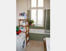 Badezimmer mit großem Fenster und beiger Fliesenboden. Zu sehen sind eine Badewanne mit grünen Mosaikfliesen, eine Dusche mit Glastür, eine Waschmaschine und ein Holzregal mit Pflegeprodukten und Handtüchern.