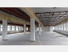 Weitläufiger, leerer Innenraum eines Gebäudes im Rohzustand mit sichtbaren Stahlträgern an der Decke, freistehenden Stützpfeilern und großen Fenstern entlang der rechten Wand. Der Boden ist betoniert, und die Wände zeigen Spuren von Verfall.