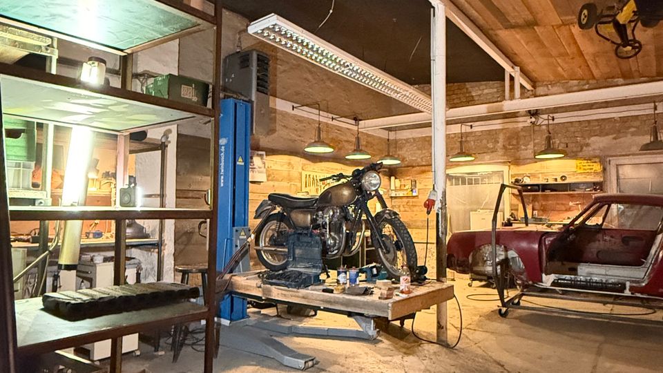Innenansicht einer Werkstattgarage mit Holzbalkendecke, Betonboden und Werkregalen an der Seite. In der Mitte steht ein Motorrad auf einer Hebebühne, im Hintergrund ein teilweise zerlegtes Auto, beleuchtet von mehreren Hängelampen.