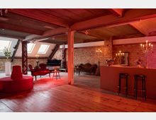 Weitläufiger loftartiger Raum mit Backsteinwänden und holzverkleideter Decke, ausgestattet mit vintage-inspirierten roten und schwarzen Sitzmöbeln. Der Raum ist stimmungsvoll beleuchtet durch Kerzenständer und hat große Dachfenster, die Tageslicht hereinlassen.