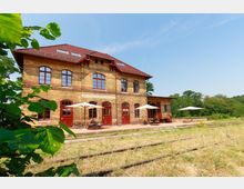Backsteingebäude eines ehemaligen Bahnhofs mit roten Fenstern und Türen, davor ein gepflasterter Bereich mit Sonnenschirmen und Tischen. Im Vordergrund sind überwachsene Gleise und Vegetation zu erkennen, im Hintergrund Bäume und blauer Himmel.