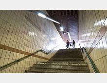 Blick auf eine Treppe in einer U-Bahn-Unterführung mit beigen, gefliesten Wänden und grünen Handläufen. Zwei Personen gehen die Treppe hinauf in Richtung eines hell erleuchteten Ausgangs.