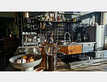 Ein Café oder private Küche mit industriellem Design, ausgestattet mit einer Espressomaschine, einem Waschbecken und einer Arbeitsfläche mit Kochutensilien. Im Hintergrund sind schwarze Kacheln, Regale mit Geschirr und Flaschen, und ein Kühlschrank sichtbar.