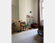 Ein Spielzimmer mit hellen Wänden und Teppichboden. Zu sehen sind ein Regal mit Kinderbüchern, ein Spielzeug-Kran, eine kleine Holz-Werkbank, ein Sitzsack und weitere Spielsachen, darunter bunte Stapelringe und ein Aufbewahrungskorb. Ein großes Fenster mit beigem Vorhang lässt Tageslicht herein.