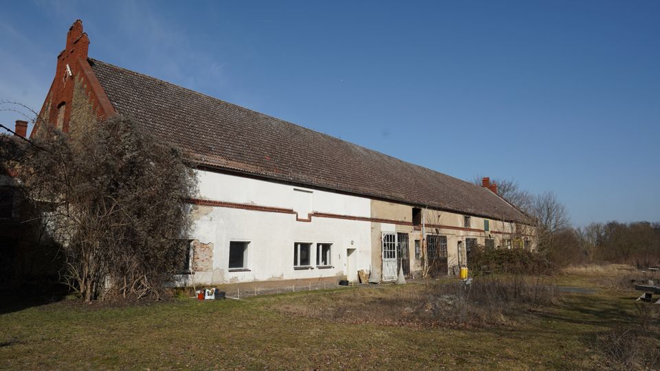 Langes, einstöckiges Wirtschaftsgebäude mit stark geneigtem Ziegeldach, teils verputzter und teils freigelegter Backsteinfassade sowie mehreren Fenstern und Toröffnungen. Davor liegt eine verwilderte Hoffläche mit Büschen und kahlen Bäumen am Rand einer ländlichen Umgebung unter klarem Himmel.