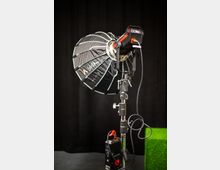 Studio-Setup mit einem professionellen Scheinwerfer, ausgestattet mit einem schwarzen Softbox-Lichtformer und montiert auf einem Metallstativ. Im Hintergrund ist ein schwarzer Vorhang und ein Teil einer grünen Fläche sichtbar.