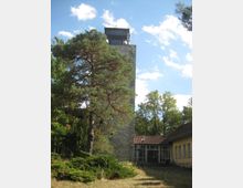 Steinerner Aussichtsturm mit quadratischem Grundriss, umgeben von Bäumen, darunter eine einzelne Kiefer im Vordergrund. Rechts befindet sich ein angrenzendes Gebäude mit gelben Wänden und einem roten Ziegeldach.