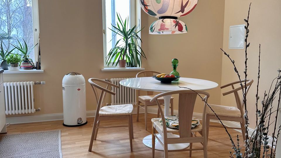 Heller Essbereich in einer Wohnung mit zwei hohen Fenstern, hellem Holzboden und runden Tisch mit vier Holzstühlen. Über dem Tisch hängt eine gemusterte Papier-Pendelleuchte; auf den Fensterbänken stehen Zimmerpflanzen, daneben ein weißer Heizkörper und ein großer Bodenvase mit Zweigen.