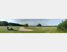 Panorama eines Golfplatzes mit einem Sandbunker in der Mitte, umgeben von gepflegten Rasenflächen und Bäumen. Links steht ein Golfcart mit Golfschlägern vor einer Baumreihe, im Hintergrund ist eine offene Wiese sichtbar.