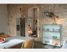 Esszimmer mit industriellem Design: Eine Wand aus rohem Backstein enthält einen großen, beleuchteten Spiegel. Links steht ein grauer Kaminofen, rechts ein offenes Regal mit Geschirr und Gläsern. Darüber hängen Töpfe an einer Metallaufhängung. Im Vordergrund ein Esstisch mit Stühlen.