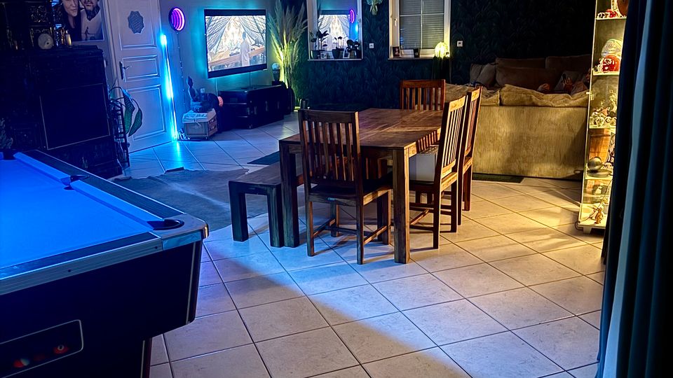 Großer Wohn- und Essbereich mit Fliesenboden, Esstisch mit Stühlen in der Mitte, Sofa-Ecke und beleuchteter Vitrine rechts. Links steht ein Billardtisch, an der Wand hängen ein Fernseher und Dekoration; farbige LED-Beleuchtung und zwei Fenster prägen den Raum.