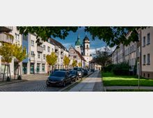 Straßenszene mit mehrstöckigen Wohnhäusern und geparkten Autos auf der linken Seite sowie einer grünen Rasenfläche rechts; im Hintergrund sind eine historische Kirche mit Spitz- und Zwiebelturm sowie ein Gebäude mit Fachwerkfassade zu sehen.