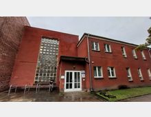 Zweistöckiges Gebäude mit roter Fassade und Flachdach, großen Glasbausteinfenstern auf der linken Seite und mehreren rechteckigen Fenstern auf der rechten Seite. Vor dem Eingang mit weißen Doppeltüren befinden sich Fahrradständer und ein gepflasterter Weg, daneben ein kleiner Grünbereich.