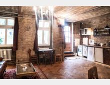 Ein rustikaler Raum mit freiliegenden Backsteinwänden, großen Fenstern und einer Holztür. Der Raum verfügt über einen Holzboden, eine Küche mit offenen Regalen und Elektrogeräten sowie eine Sitzecke mit einem kleinen Tisch und Stühlen.