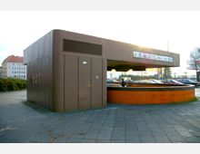 Brauner, moderner U-Bahn-Eingang mit rechteckiger Überdachung und orange gekacheltem Geländer, umgeben von einer gepflasterten Fläche und parkenden Autos im Hintergrund. Oben auf dem Dach sind Richtungsschilder mit Hinweisen zu nahegelegenen Zielen wie Messe und ZOB angebracht.