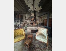 Ein rustikaler Raum mit einer alten, abgenutzten Backsteinwand, einem Kamin in der Mitte und zahlreichen Werkzeugen an den Wänden. Im Vordergrund stehen zwei gepolsterte Sessel im Vintage-Stil, kleine Holzhocker und ein orientalischer Teppich auf einem rustikalen Holzboden.