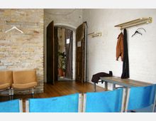 Raum mit hellen Ziegelwänden und Holzboden, ausgestattet mit blauen Kunststoffstühlen, einer Sitzbank, sowie Wandgarderoben mit Kleiderbügeln und hängender Kleidung. Im Hintergrund eine geöffnete Holztür, die den Blick in einen weiteren Raum mit Topfpflanzen freigibt.