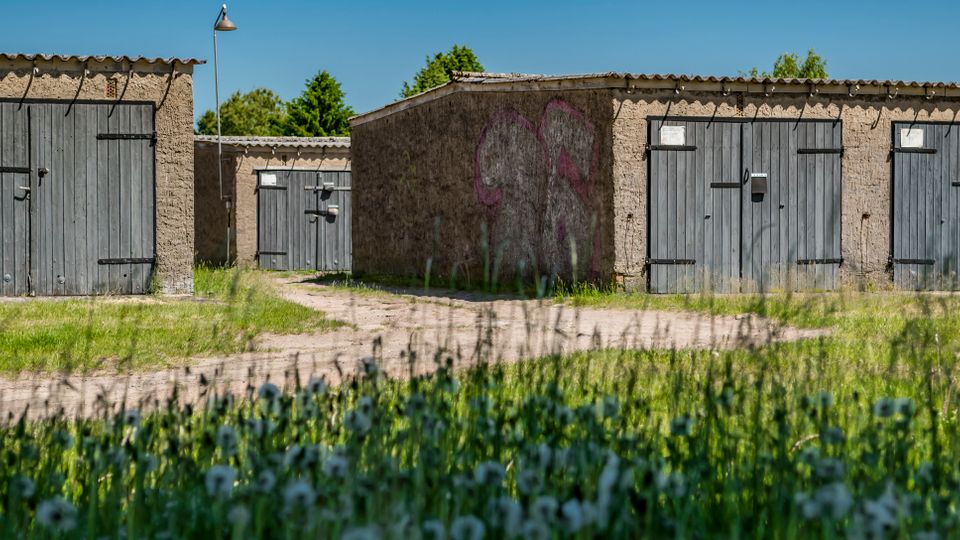 Reihe niedriger Garagengebäude aus rauem Beton mit mehreren grauen Holztoren und einem unbefestigten Weg dazwischen. Im Vordergrund liegt eine grasige Wiese mit Wildblumen, dahinter ist ein blauer Himmel zu sehen.